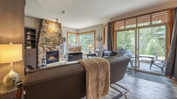 A living room with a stone fireplace, a sofa, a television, and a view of the outdoors through large windows.