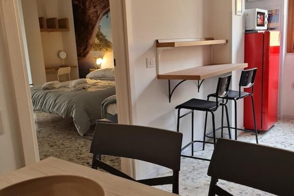 Une chambre d’hôtel de taille modeste, comprenant un lit, un petit bureau, deux chaises et un réfrigérateur rouge.