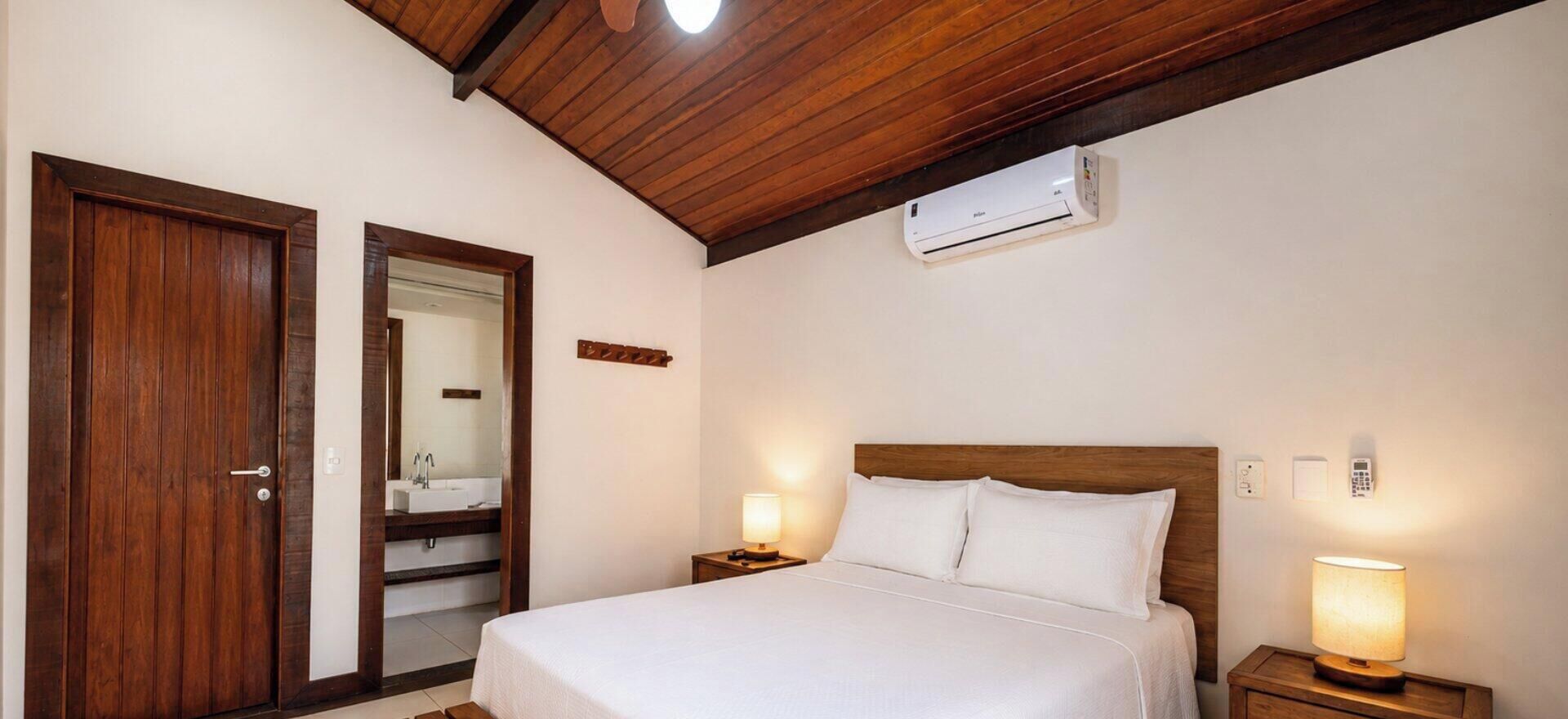 A hotel room with a wooden ceiling, a bed with white bedding, bedside lamps, a wall-mounted air conditioner, and a door leading to a bathroom.