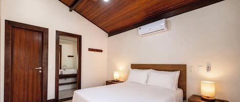 Una habitación de hotel con techo de madera, cama con ropa de cama blanca, lámparas de noche, aire acondicionado instalado en la pared y una puerta que da a un baño.