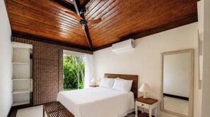 Une chambre avec un plafond en bois, un lit recouvert de draps blancs, une table de chevet avec une lampe et une fenêtre donnant sur un paysage verdoyant.