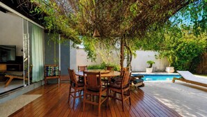 Eine Terrasse im Freien mit einem Holztisch und Stühlen, umgeben von üppigem Grün und einem Swimmingpool.