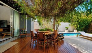 Un patio al aire libre con una mesa y sillas de madera, rodeado de abundante follaje y una piscina.
