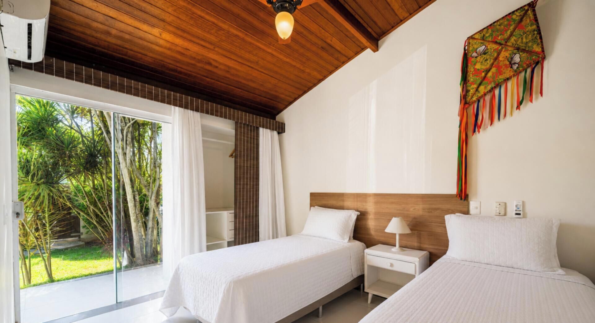 A hotel room with two beds, a wooden ceiling, a large window with curtains, and a view of greenery outside.