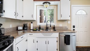 Una cocina con armarios blancos, encimera de granito, estufa, lavavajillas y ventana con vistas a árboles.