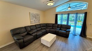 A living room with a sectional sofa, a white coffee table, a ceiling fan, and a view of a swimming pool through large windows.