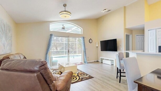 A spacious living room with a large window, a ceiling fan, a flat-screen TV, and a dining area with a table and chairs.