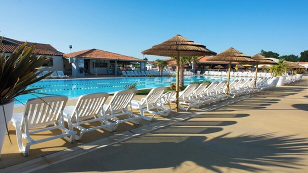 Un espace aménagé au bord de la piscine, avec des chaises longues et des parasols, une piscine à l’eau d’un bleu limpide, et des bâtiments aux toits couverts de tuiles en arrière-plan.