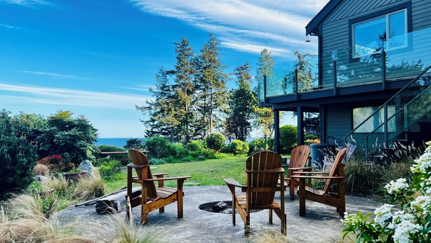 Une terrasse dotée de chaises en bois et d’un foyer, offrant une vue sur un jardin luxuriant et une maison aux grandes fenêtres.