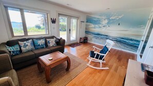 Un salon doté d’un plancher en bois, d’un canapé agrémenté de coussins aux motifs marins, d’un fauteuil à bascule et d’une fresque représentant l’océan.