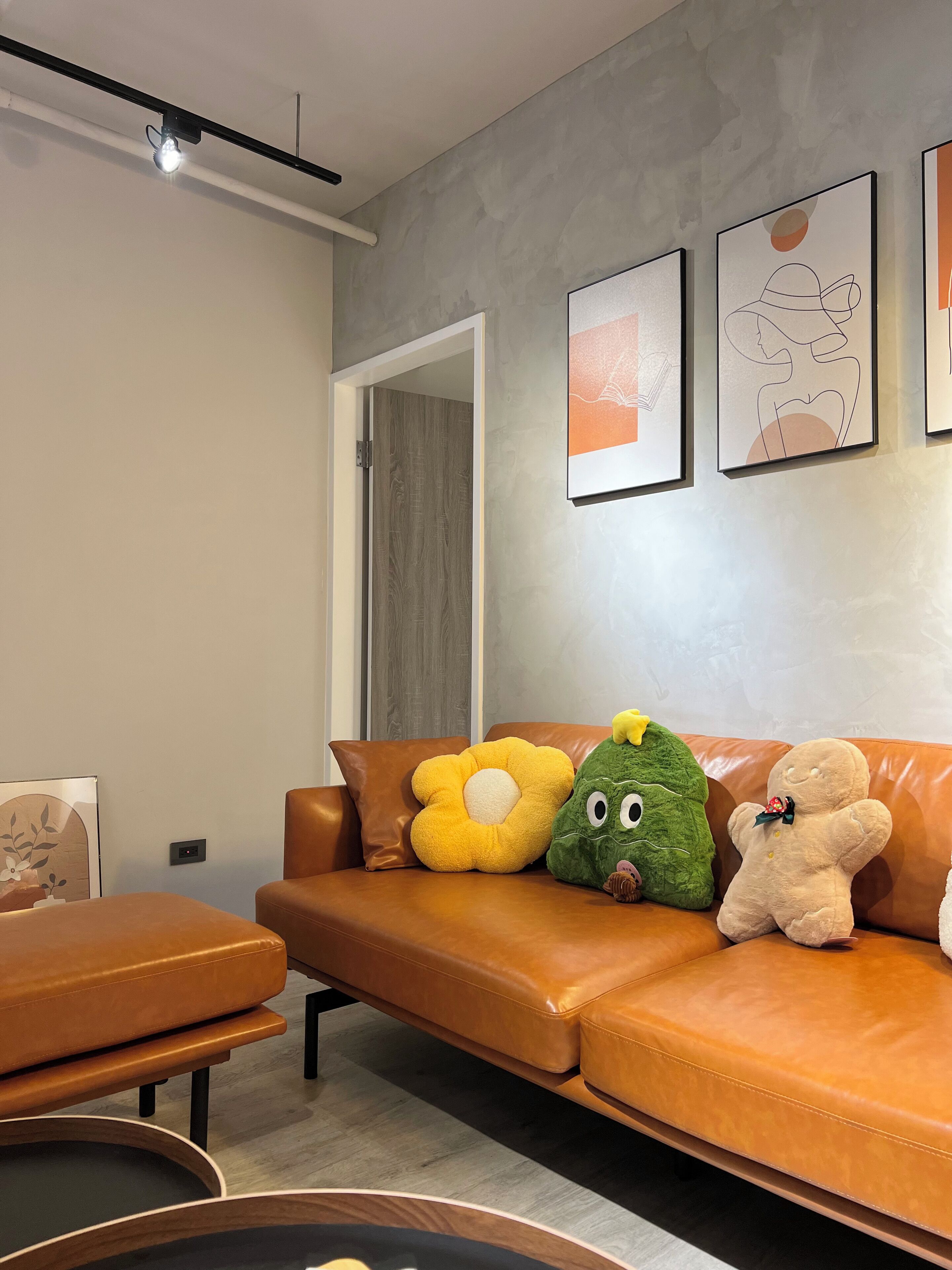 A modern living room with a brown leather sofa, a small round table, and a wooden coffee table. There are three framed artworks on the wall and a door leading to another room.