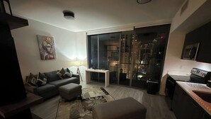 A modern living room with a sofa, ottoman, and a coffee table. There is a television, a lamp, and a painting on the wall. A sliding glass door leads to a balcony with a city view. - Miami Downtown 27 floor Luxe Escape rooftop pool and jacuzzi (Miami)