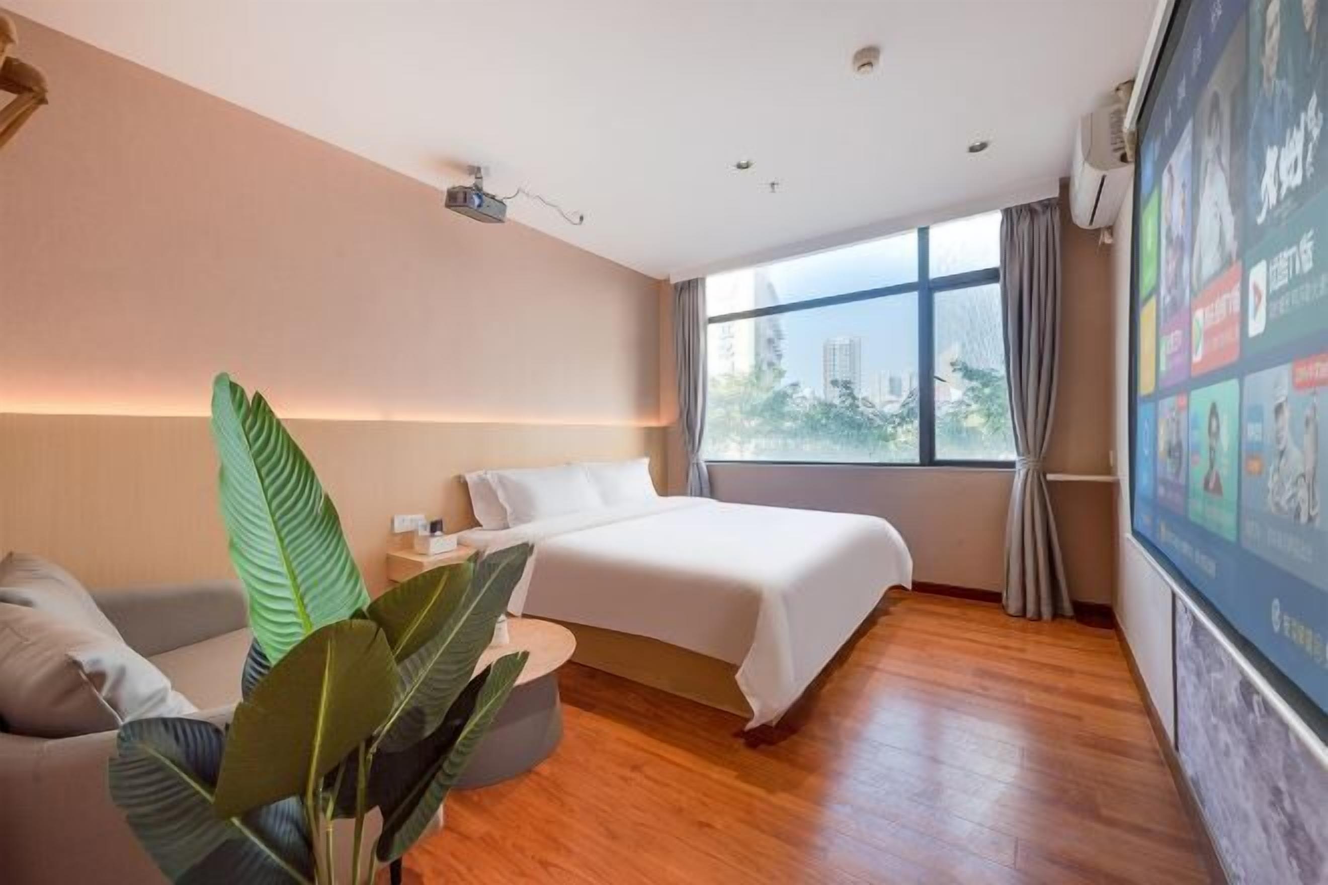 Een hotelkamer met een bed, een bank, een plant, een projector en een raam met uitzicht op de stad.