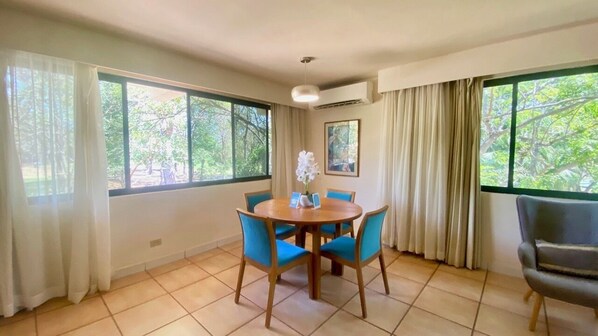 Un comedor con una mesa redonda y cuatro sillas, ventanas amplias con cortinas y vistas a zonas verdes.
