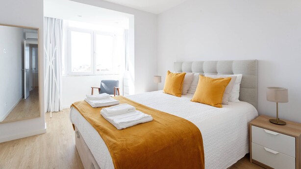 A modern hotel room with a large bed, a nightstand, a lamp, and a view of the city through the window.