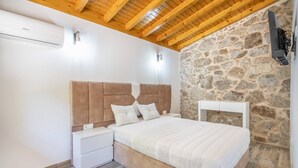Une chambre avec un mur en pierre, des poutres apparentes en bois au plafond, un lit, une table de chevet et un téléviseur fixé au mur.