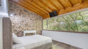 Un dormitorio con pared de piedra, techo de madera y un ventanal con vistas a árboles.