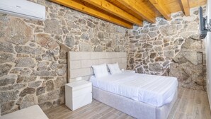 A bedroom with a stone wall, wooden ceiling beams, a bed, a nightstand, and a white air conditioning unit. - Casa do Penedo - Aqua View (Vila Verde)