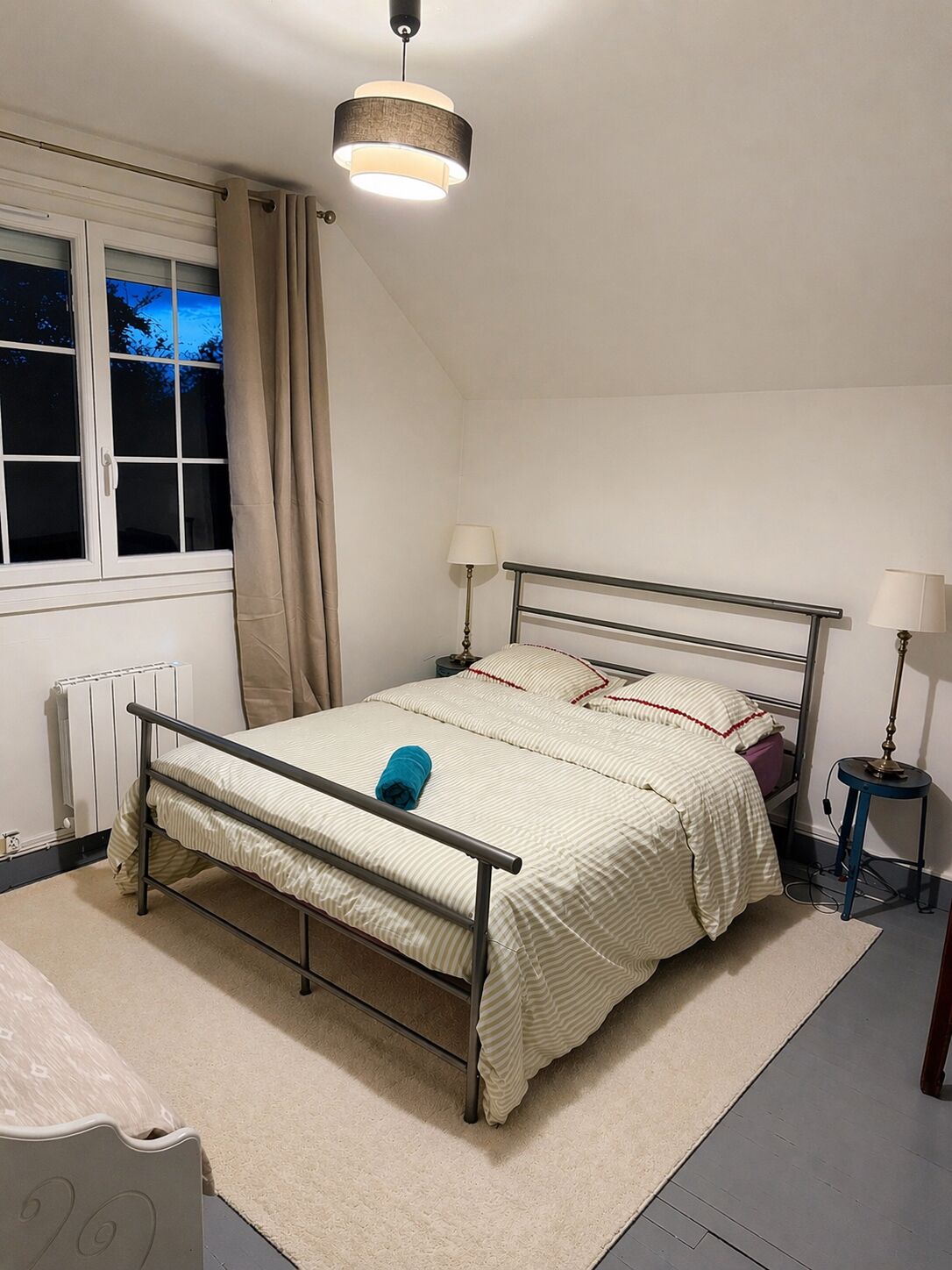 A bedroom with a bed, bedside lamps, a radiator, a window with curtains, and a hanging light fixture.