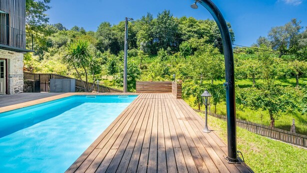 A wooden deck with a swimming pool, surrounded by lush greenery and a stone building.