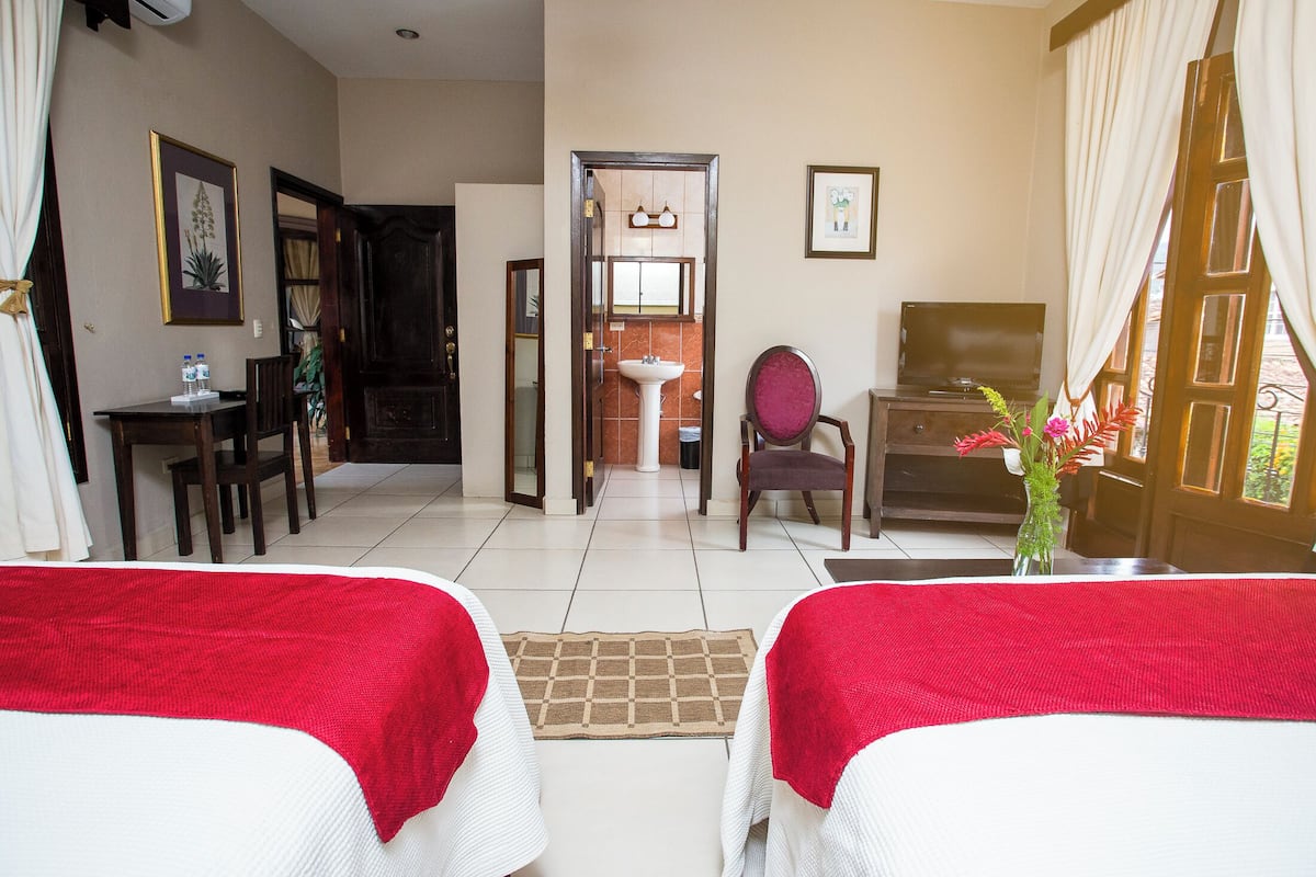 A hotel room with two beds, a desk, a chair, a TV, and a bathroom visible through an open door.