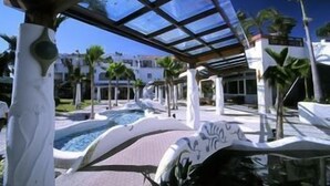Zona de piscina con techo de vidrio, palmeras y un edificio al fondo.