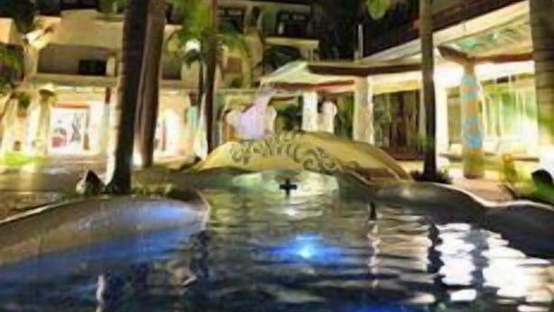 Vista nocturna de un resort con piscina, palmeras y edificios iluminados.