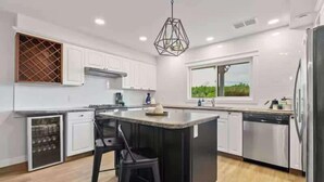 A modern kitchen with white cabinetry, a central island with bar stools, a built-in wine cooler, and a view of greenery outside.