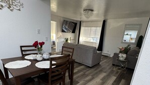 A modern living room with a grey sofa, a dining table with chairs, and a TV mounted on the wall.