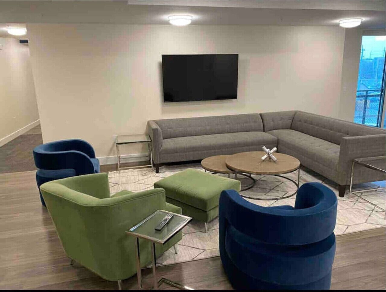 A modern living room with a grey sectional sofa, green and blue armchairs, a round coffee table, and a flat-screen TV mounted on the wall.