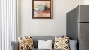 Ein Wohnzimmer mit grauem Sofa, gemusterten Kissen, einem gerahmten abstrakten Gemälde und einem Edelstahl-Kühlschrank.