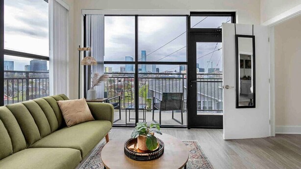 A modern living room with a green sofa, a wooden coffee table with a plant, and a view of a cityscape through large sliding glass doors. - Modern Penthouse Condo with Panoramic City View (Nashville)