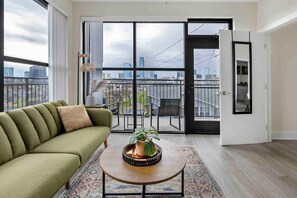 Een moderne woonkamer met een groene bank, een houten salontafel met een plant, en een stadsgezicht door grote schuifpuien.