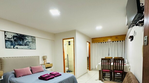 Une chambre d’hôtel comprenant un lit, un banc, une table à manger avec des chaises, un téléviseur fixé au mur et un tableau accroché au mur.