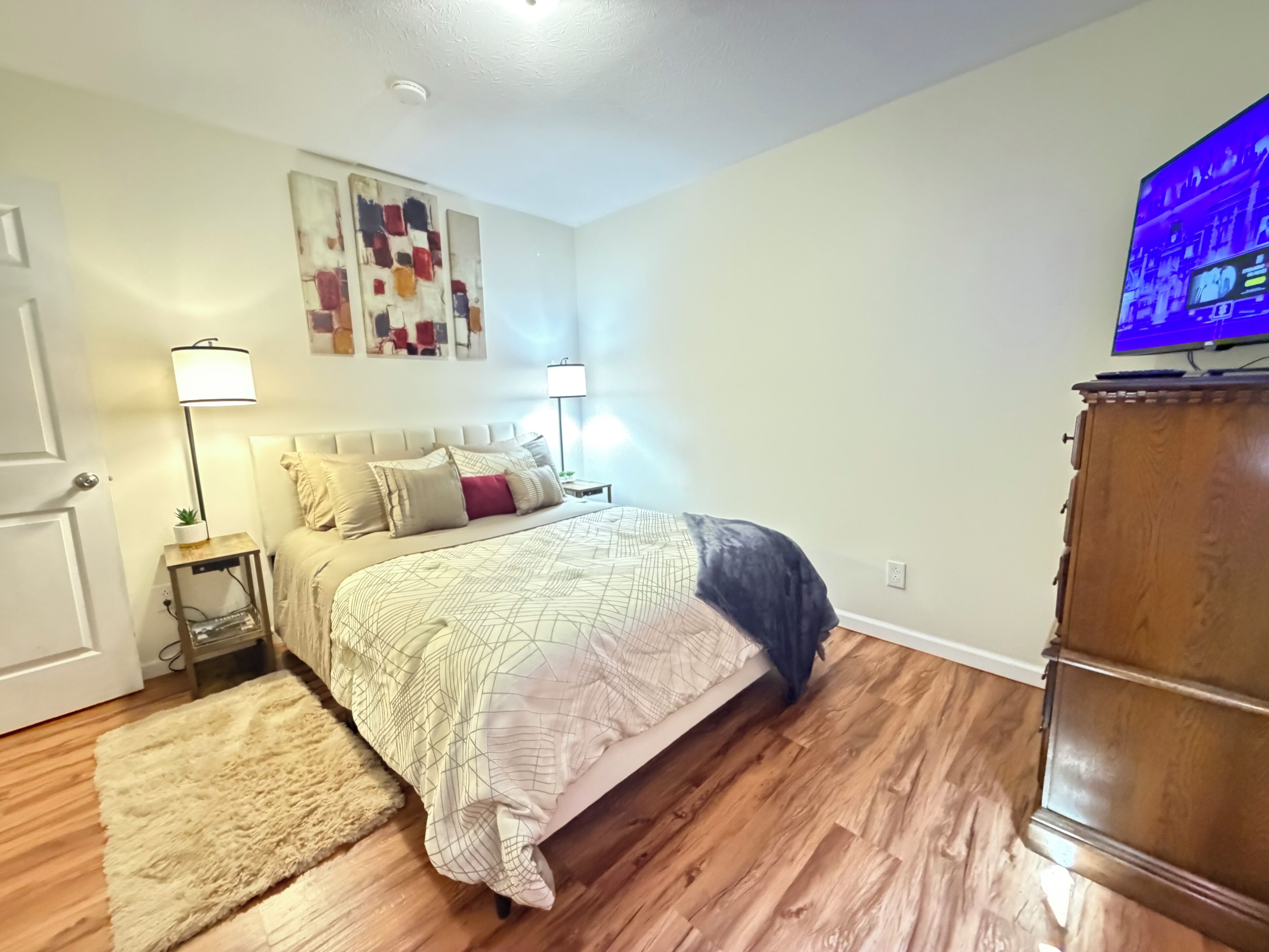 A bedroom with a bed, two bedside lamps, a wooden floor, a TV on a wooden cabinet, and a door.