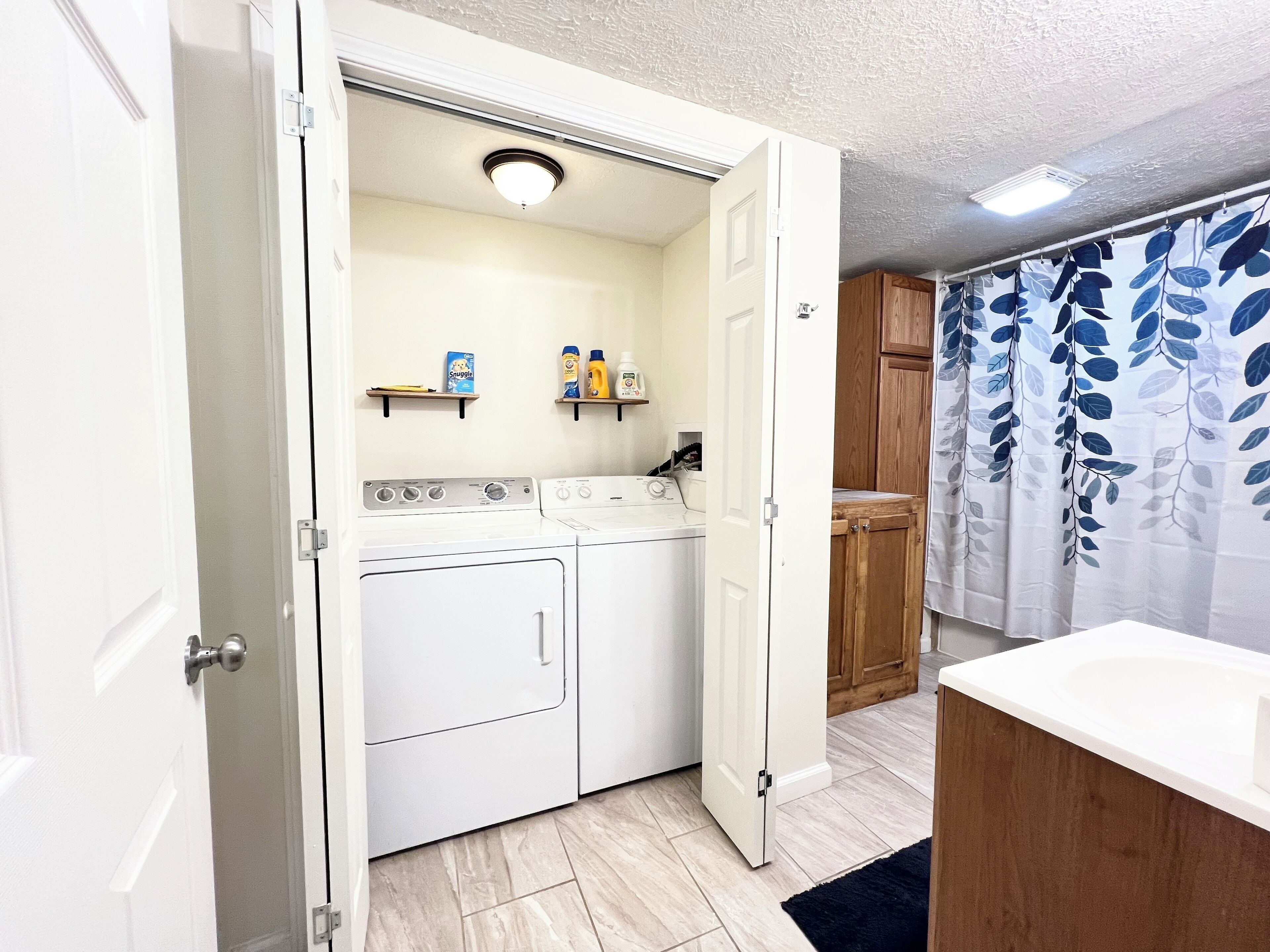 A laundry room with a washing machine and dryer, wooden cabinets, and a shower area with a leaf-patterned curtain.