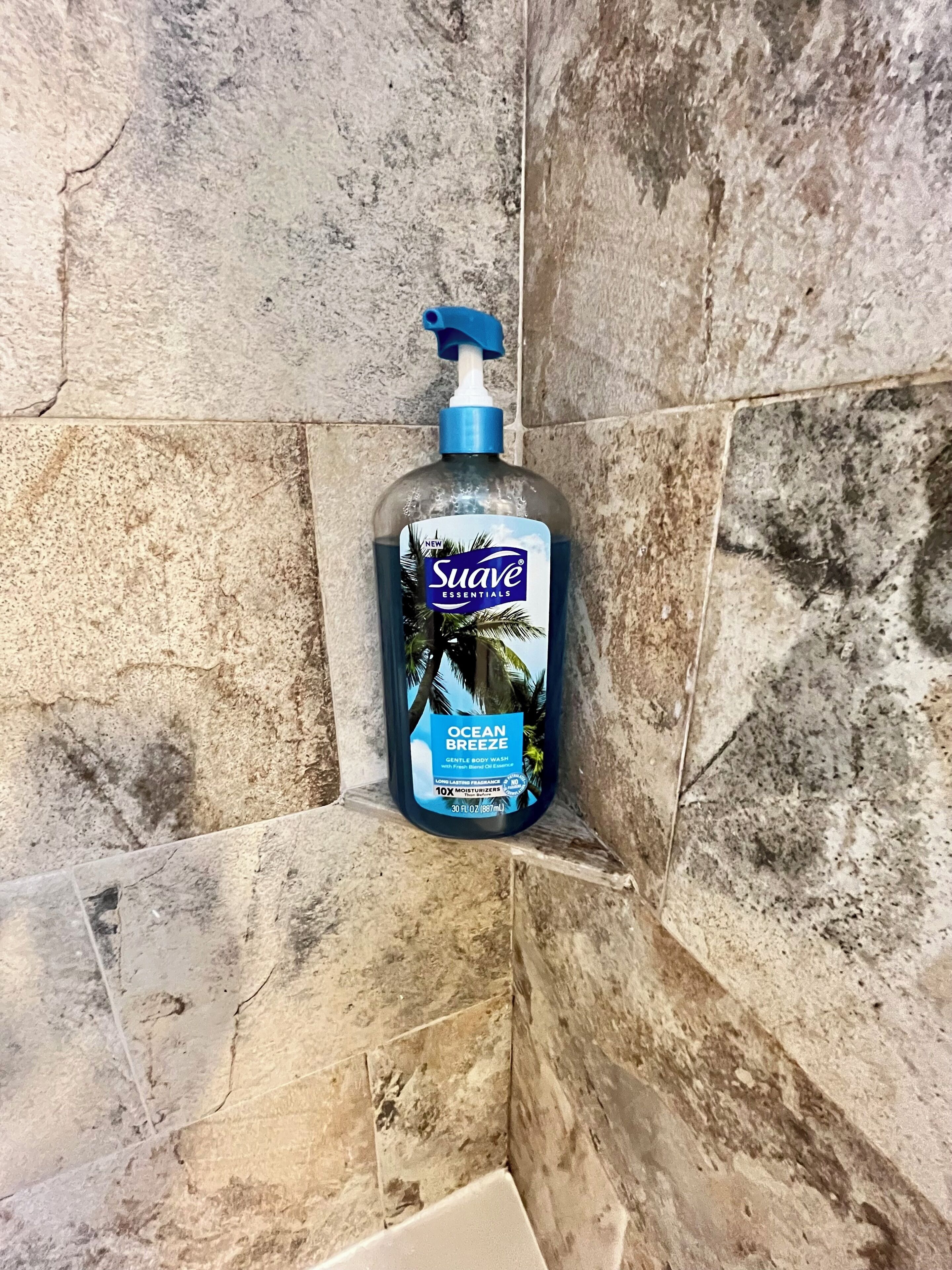 Um frasco de Suave Essentials Ocean Breeze para o corpo em um canto de um banheiro com azulejos de pedra.