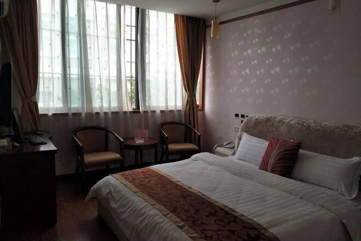 A hotel room with a bed, two chairs, a desk with a computer, and a window with curtains.