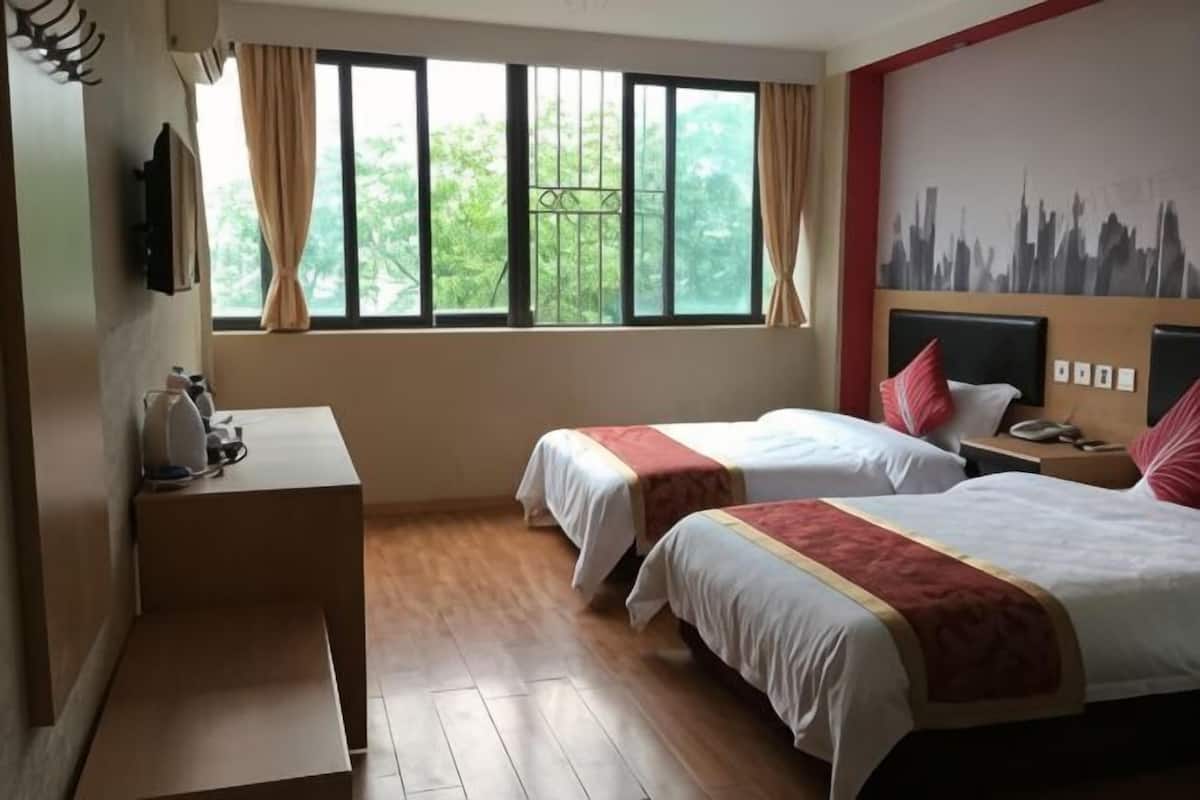 A hotel room with two beds, a desk, a chair, a window with curtains, and a cityscape mural on the wall.