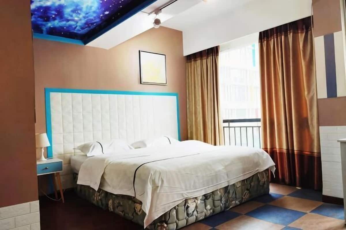 A hotel room with a bed, a nightstand, a window with curtains, and a wall with a painted design.