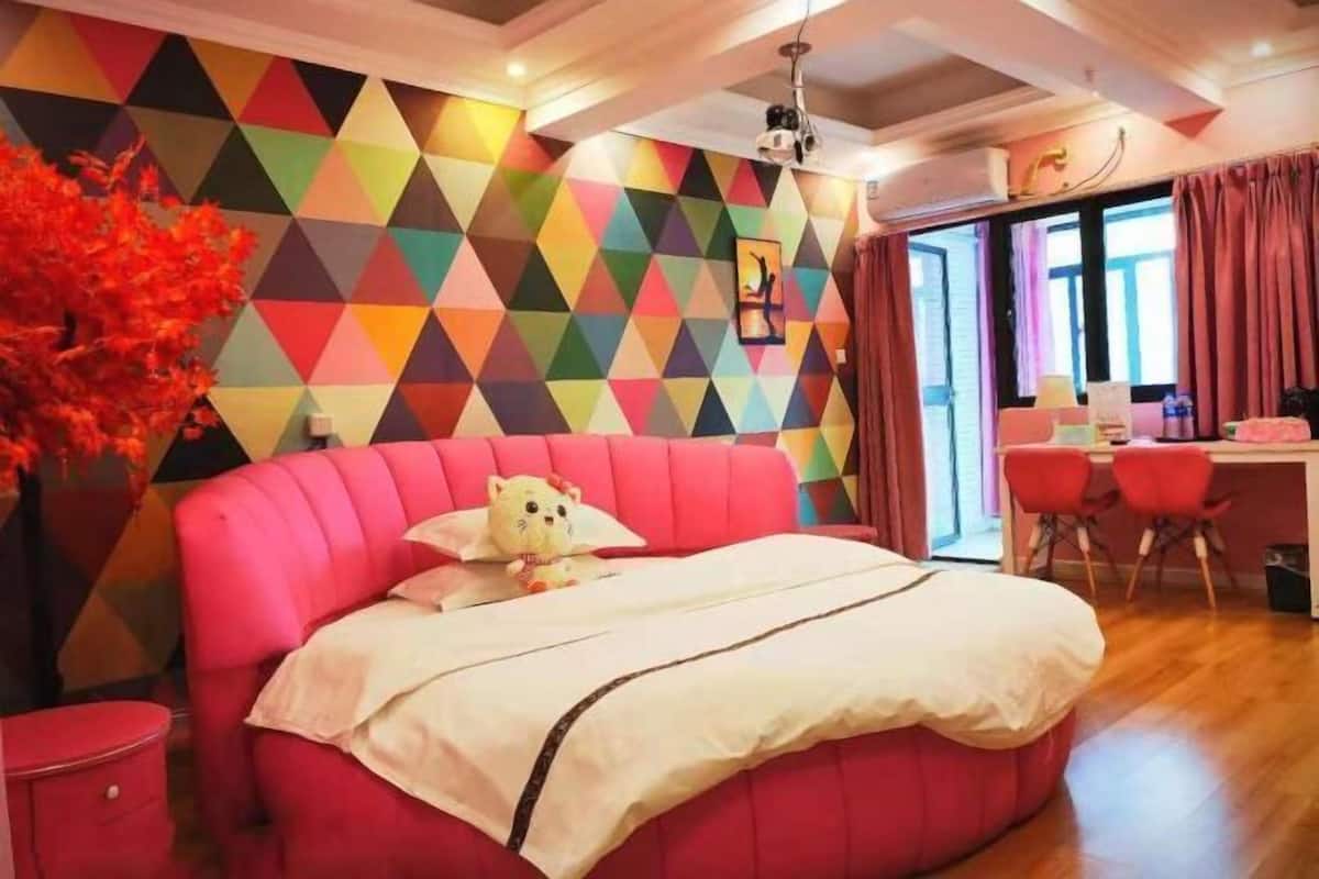 A modern bedroom with a vibrant geometric wall, a plush red bed, and a desk with red chairs.