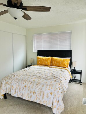 Un dormitorio con cama, ventilador de techo, ventana con persianas y mesita de noche.