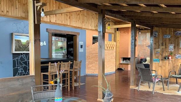 An outdoor seating area with wicker chairs and a glass table, wooden beams overhead, and a brick wall with various signs. - Pousada Deck 950 (Cambará do Sul)
