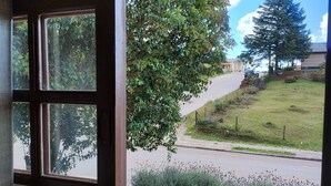 A window with a view of a road, greenery, and a building in the distance. - Pousada Deck 950 (Cambará do Sul)