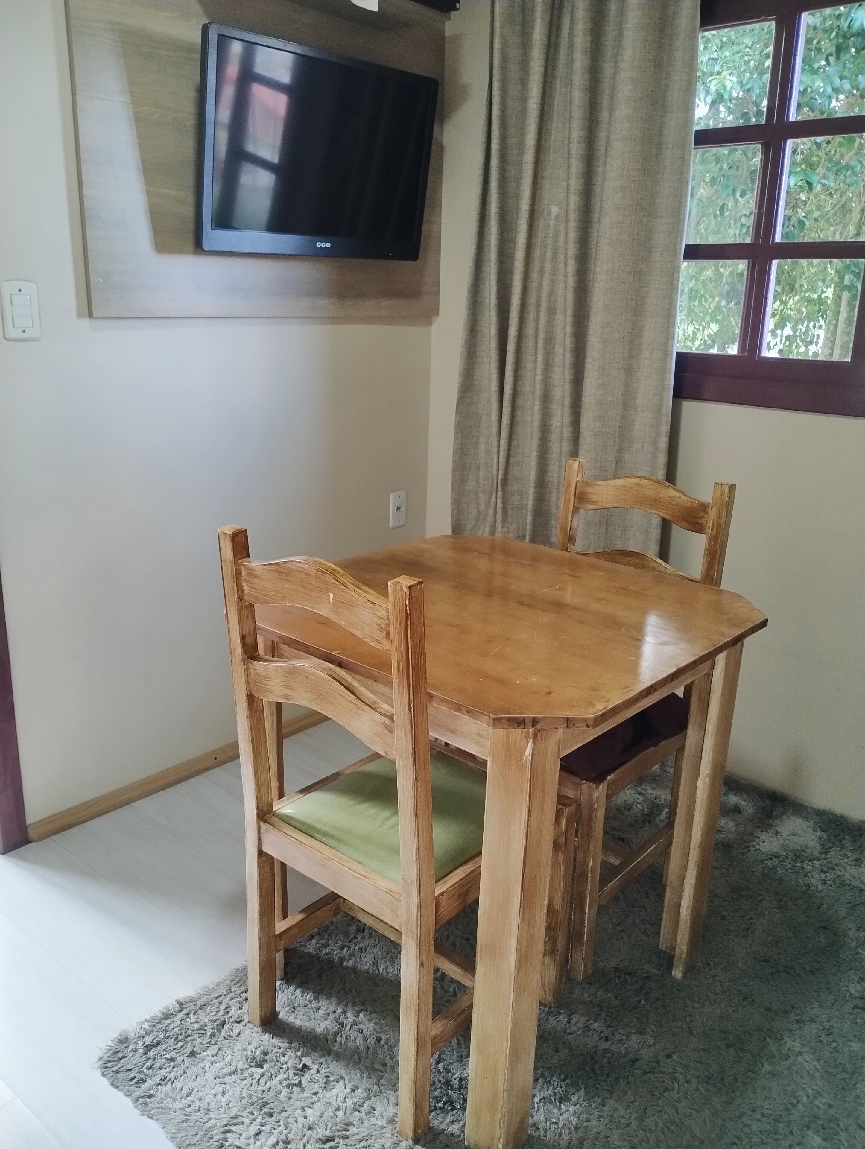 Una mesa de madera con cuatro sillas, un televisor montado en la pared y una ventana con cortinas.