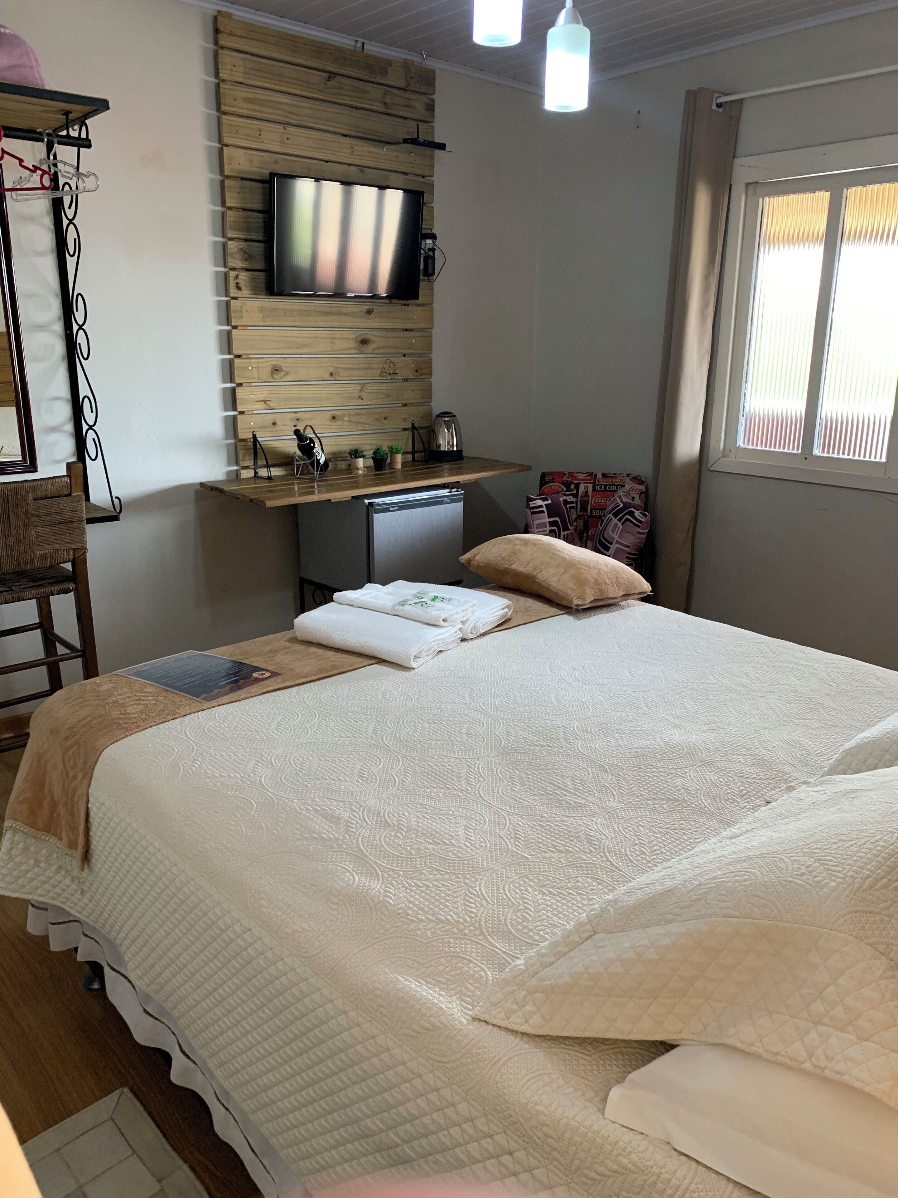 Un dormitorio con cama, una kitchenette pequeña, un televisor montado en una pared de madera y una ventana con persianas.
