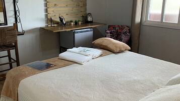 Un dormitorio con cama, una kitchenette pequeña, un televisor montado en una pared de madera y una ventana con persianas.