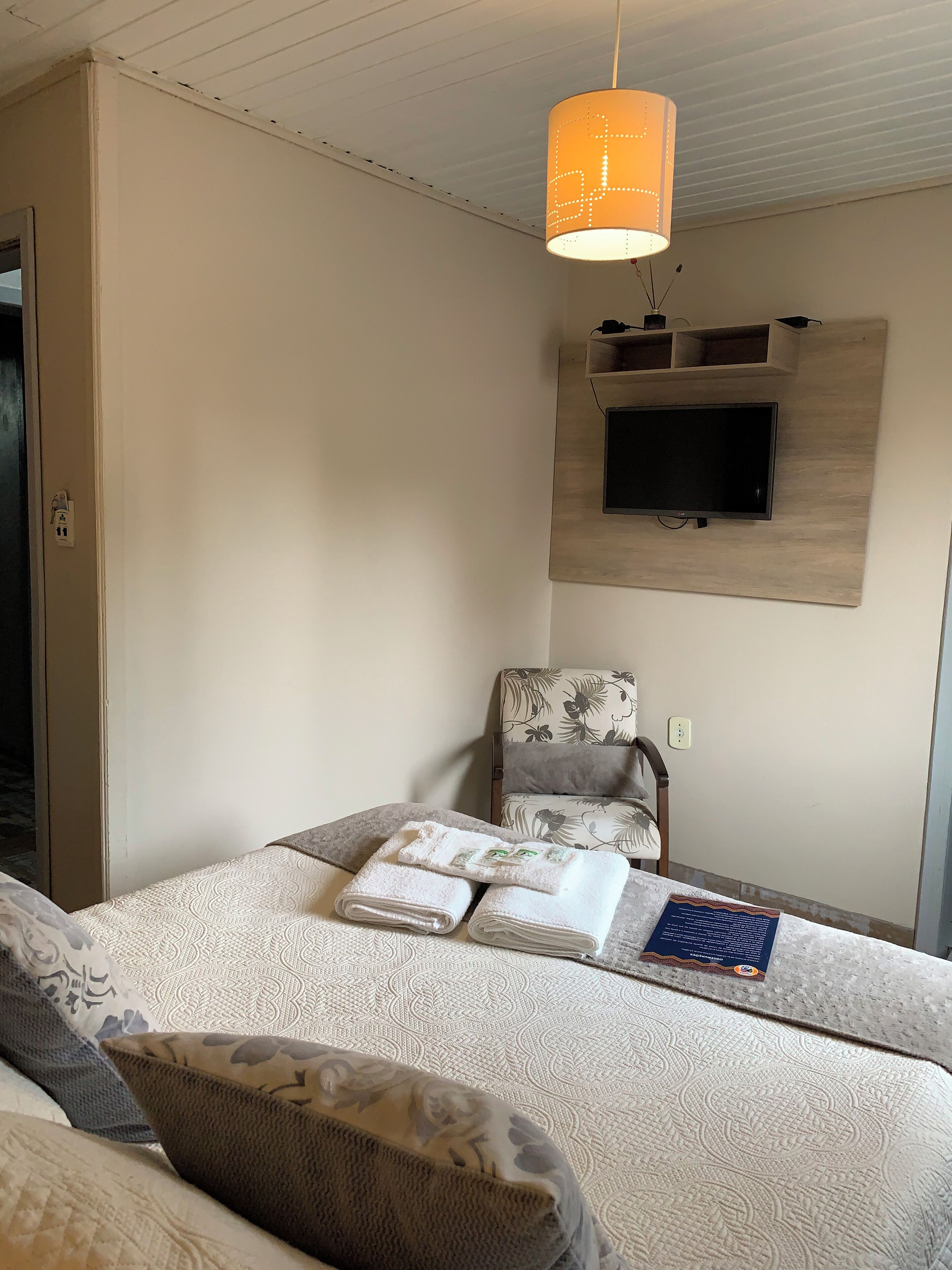 A neatly arranged bedroom with a bed, a chair, a television mounted on the wall, and a hanging light fixture.