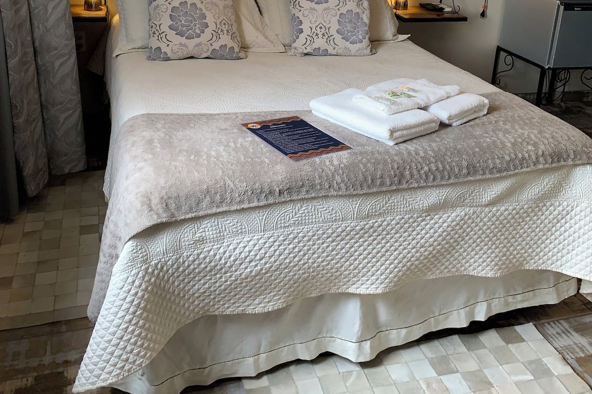 A neatly made bed with a quilted cover, a bedside table with a lamp, and a small table with a kettle and a remote control.