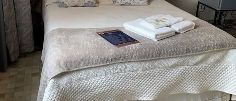 A neatly made bed with a quilted cover, a bedside table with a lamp, and a small table with a kettle and a remote control.